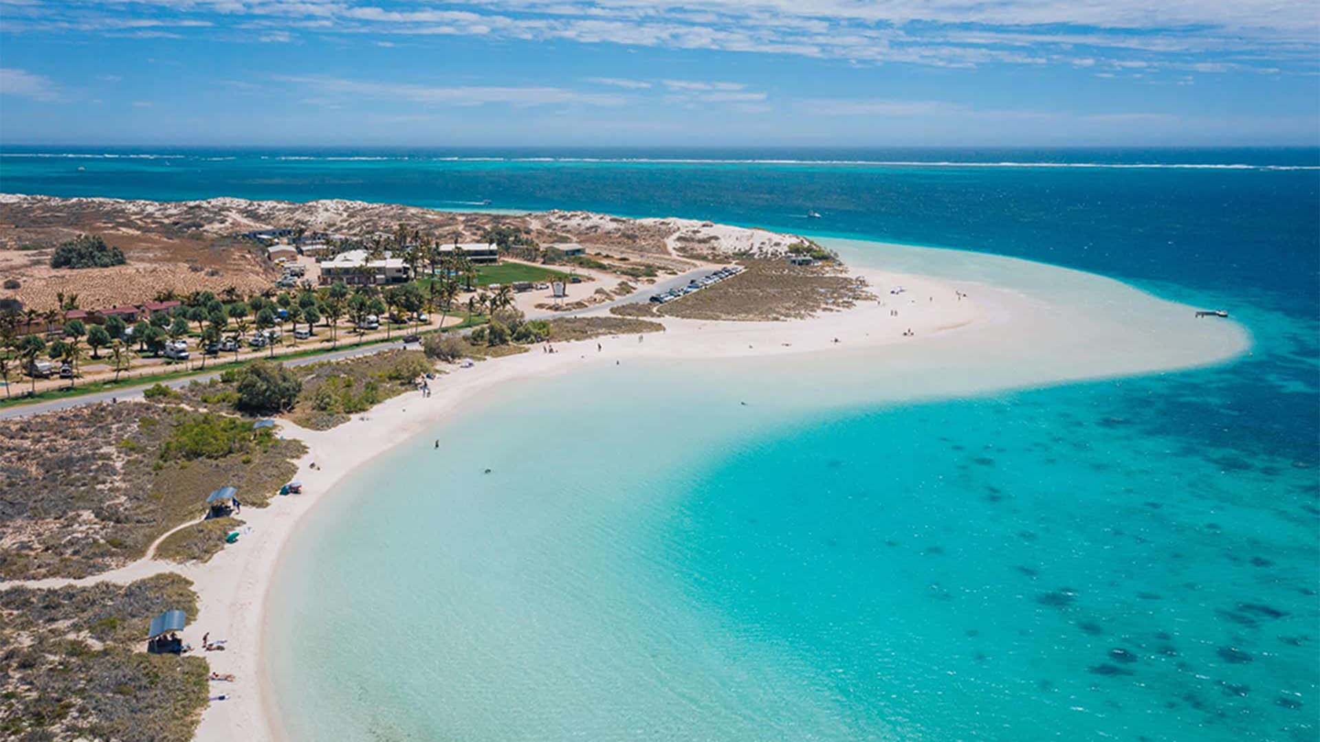 Aerial view of a turquoise bay with clear water, white sandy beaches, and sparse vegetation. Small buildings and palm trees line the shore under a bright blue sky, capturing the beauty of Paphos beaches.