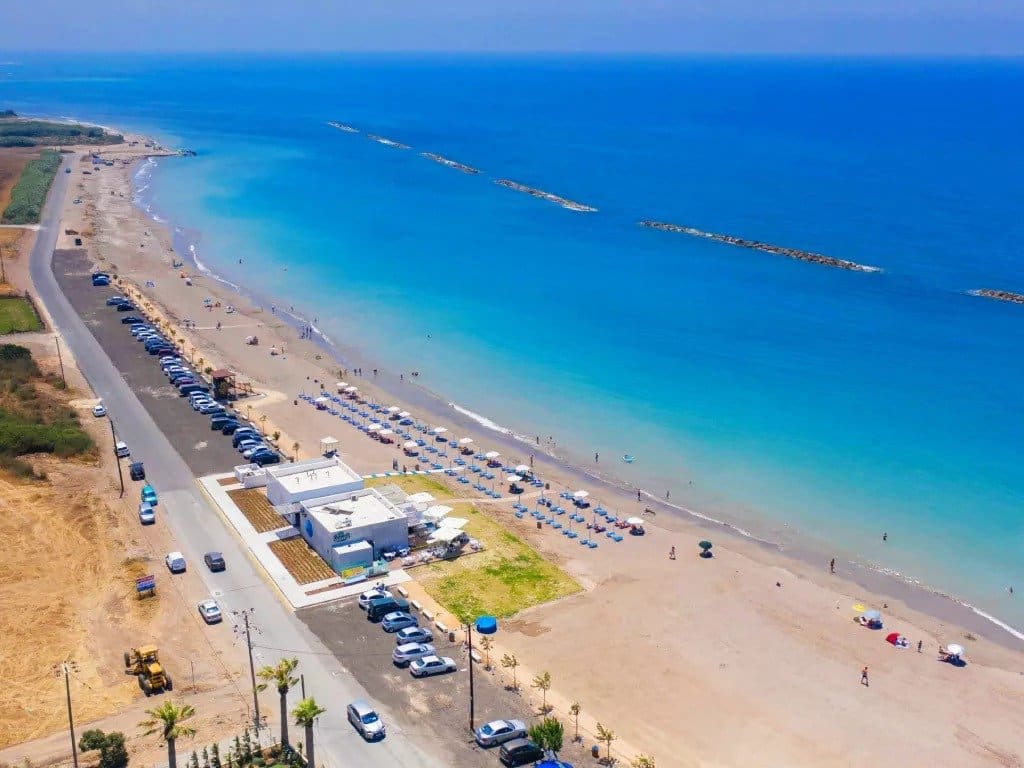 Aerial view of Paphos beaches with turquoise water, a line of umbrellas and sunbeds, parked cars along a road, and a white building near the shore. People are walking and swimming along the sandy coastline.