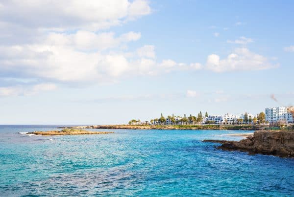 A coastal scene with clear blue water, rocky shoreline, and white buildings with palm trees in the distance under a partly cloudy sky.