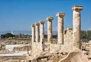 Ancient stone columns stand in a row among ruins under a clear blue sky at the Archaeological Park of Kato Pafos, with distant trees and buildings visible on the horizon.
