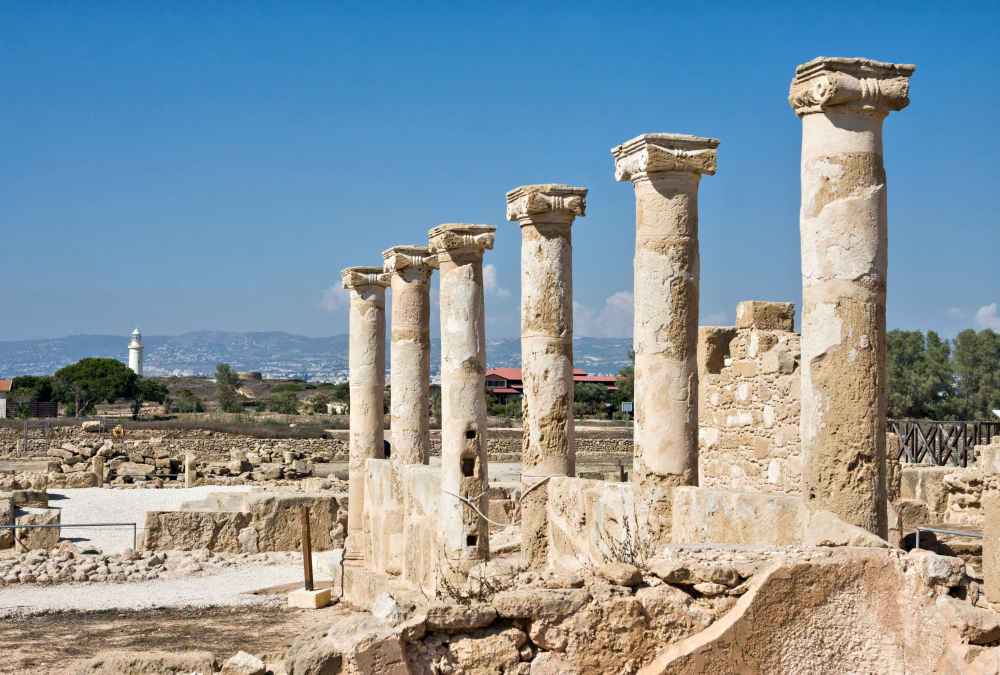 Ancient stone columns stand in a row among ruins under a clear blue sky at the Archaeological Park of Kato Pafos, with distant trees and buildings visible on the horizon.