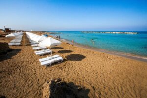 A sandy beach with rows of empty white sun loungers and parasols, calm turquoise water, and a few people swimming and playing near the shore under a clear blue sky—an inviting scene typical of Paphos beaches.