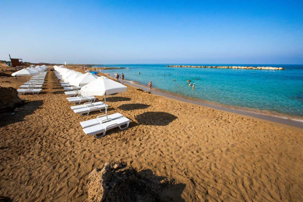 A sandy beach with rows of empty white sun loungers and parasols, calm turquoise water, and a few people swimming and playing near the shore under a clear blue sky—an inviting scene typical of Paphos beaches.