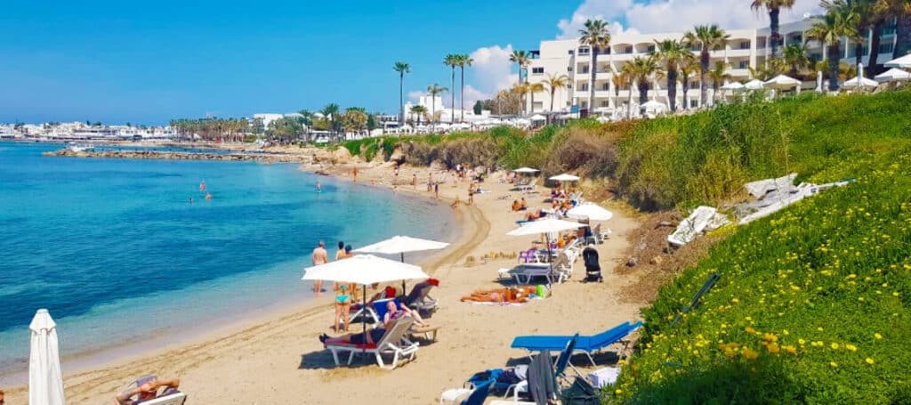 People relax on sun loungers under white parasols on a sandy beach by clear blue water. Palm trees and a white resort hotel overlook the scene, capturing the laid-back charm of Paphos beaches under a bright, sunny sky.